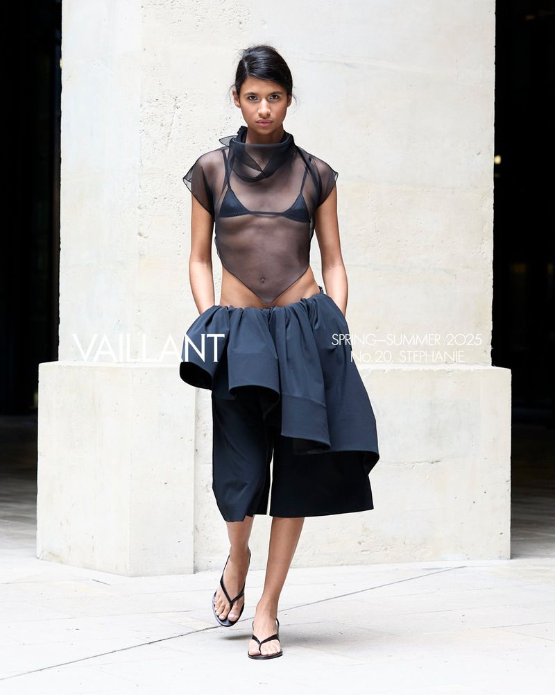 Runway fashion show featuring a model wearing a black mesh top, black bottom, and flip flops, walking in front of a concrete wall.