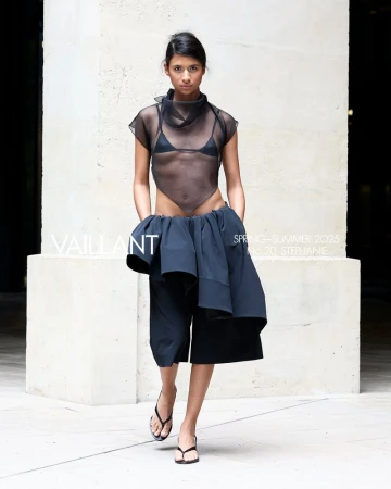 Runway fashion show featuring a model wearing a black mesh top, black bottom, and flip flops, walking in front of a concrete wall.