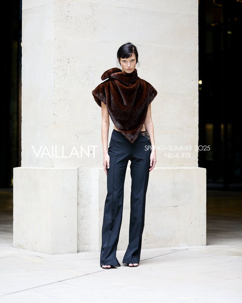Brunette woman wearing a fur top and black pants, posing against a light stone wall outdoors. Fashion editorial shot.