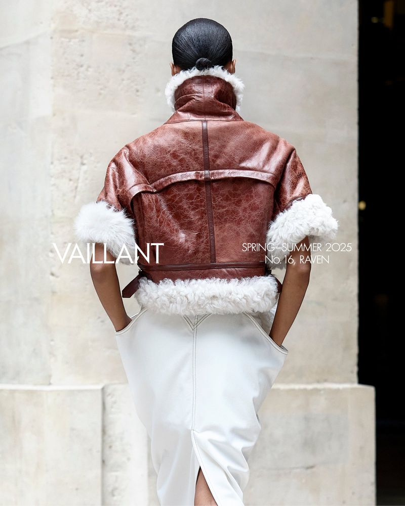 Fashion shot from behind: A model wears a brown shearling jacket with white accents and a white skirt against a stone backdrop.