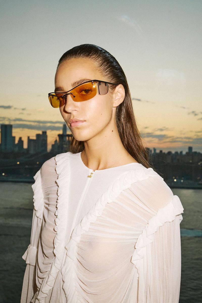 Brunette woman in sheer white dress and sunglasses poses against a city skyline at dusk. Fashion photography in an urban setting.