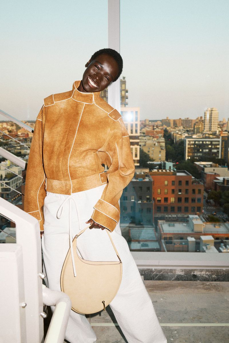 Smiling Black woman in a tan jacket and white pants stands on a balcony overlooking a cityscape, carrying a light-colored handbag.
