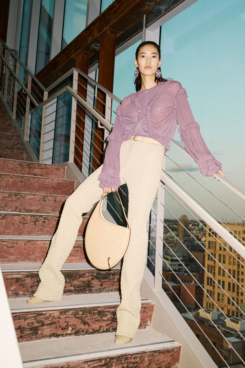 Asian woman in lilac blouse and beige pants poses on staircase with purse and platform heels, backlit by city view at dusk.