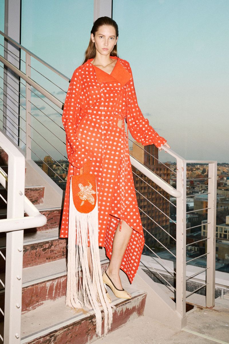 A woman in a dotted red dress and heels poses on stairs with a fringed purse in an urban setting against a sunset sky.