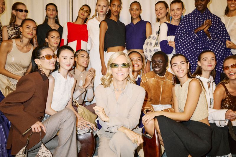 Large group of diverse female fashion models and designer posing together indoors, wearing sunglasses and various outfits. 