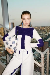 Model wearing a white and purple patterned top holding a printed handbag on a staircase with city views
