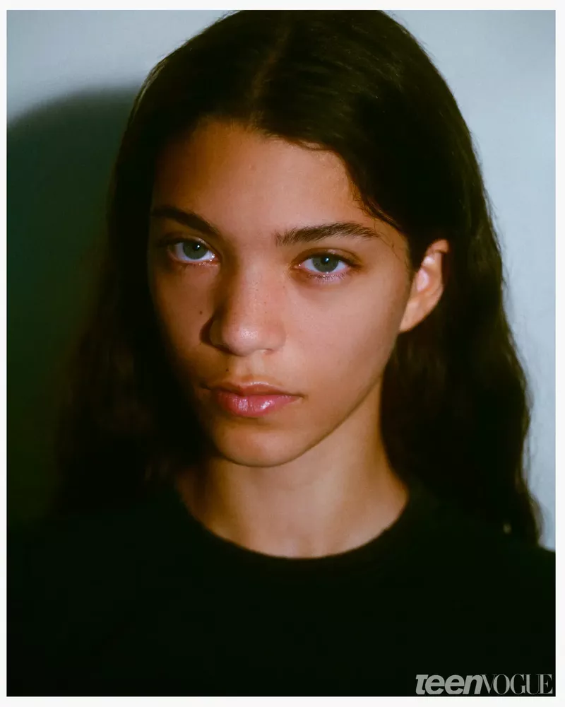 Teen Vogue headshot: A young woman with dark hair and light eyes poses indoors in a black top against a mottled white and green background.