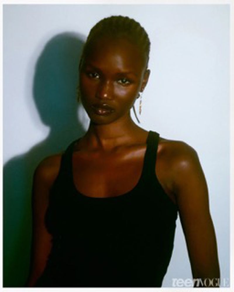 Close-up portrait of a Black female model wearing a black tank top, posed in front of a white wall with a shadow. Teen Vogue logo visible.