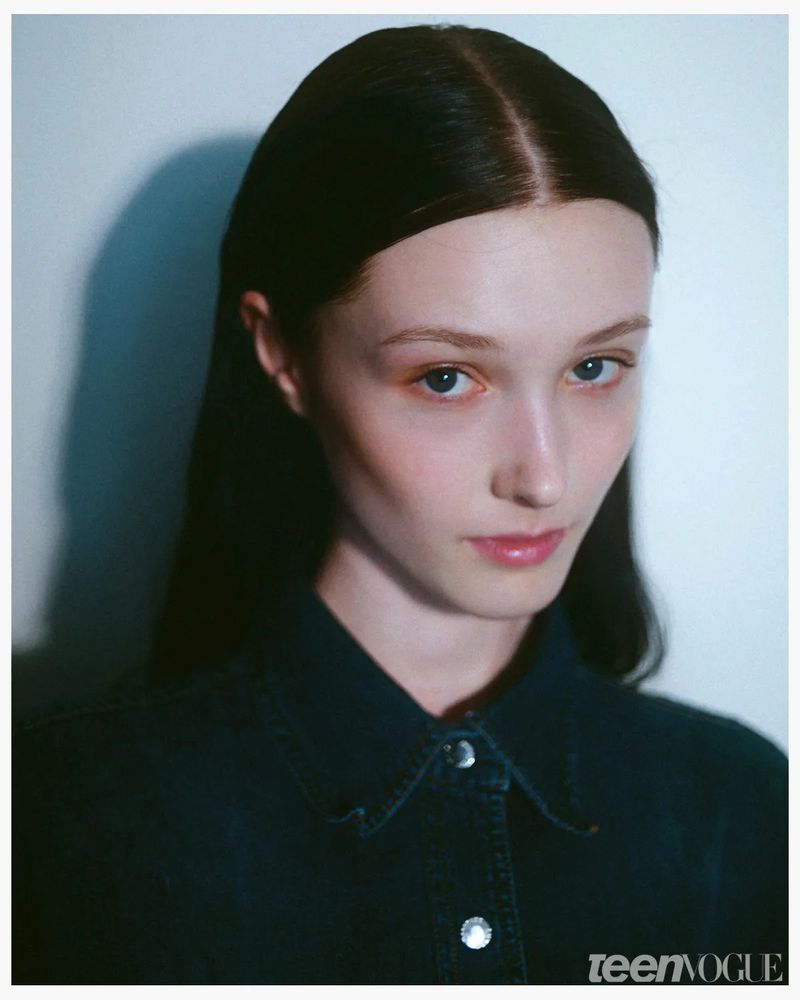 Close-up portrait of a young woman with dark hair and a denim shirt, against a soft, shadowed background. Beauty and fashion editorial.