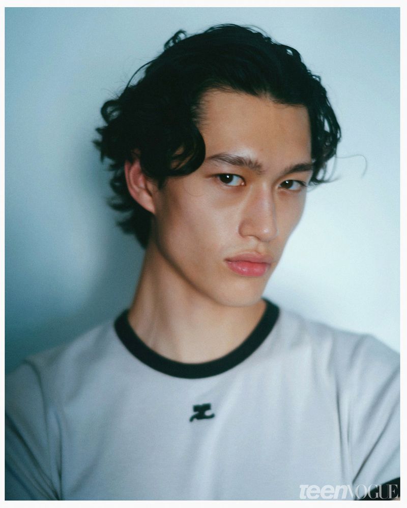 Portrait of a young Asian male model with wavy black hair, wearing a light gray t-shirt in a photo studio against a soft blue background.