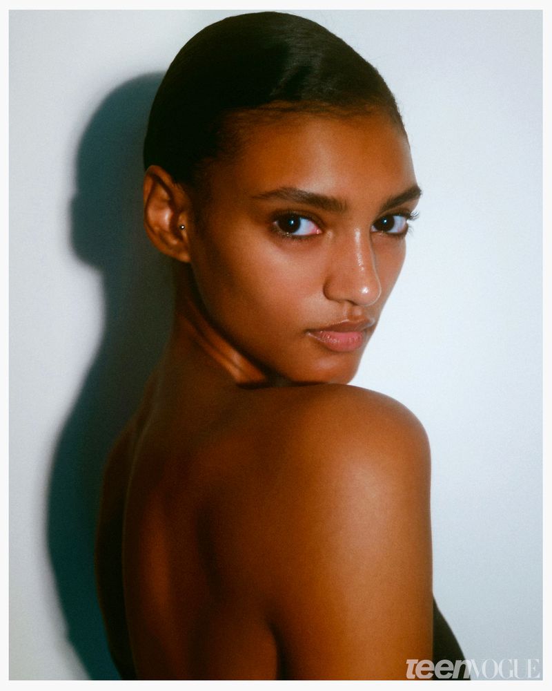 Close-up portrait of a woman with dark brown skin and slicked back hair posing in front of a white wall in a photo studio.