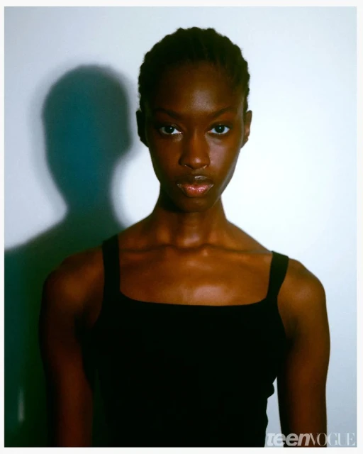 Portrait of a Black woman wearing a black tank top against a white wall with a distinct shadow cast behind her.