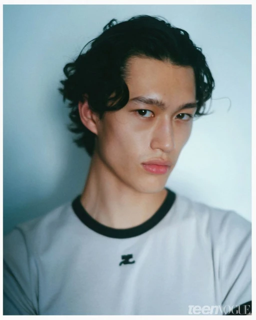 Portrait of a man with dark curly hair wearing a white t-shirt with a black collar against a light blue background.