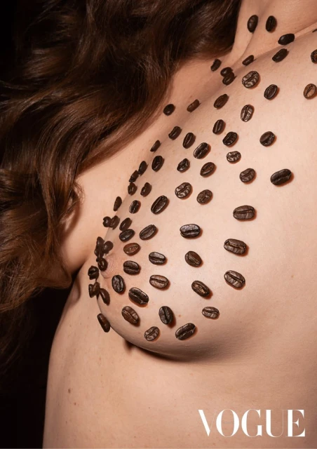Woman's nude torso covered in coffee beans, close-up shot in dark studio with Vogue logo in corner, brown hair and skin visible.
