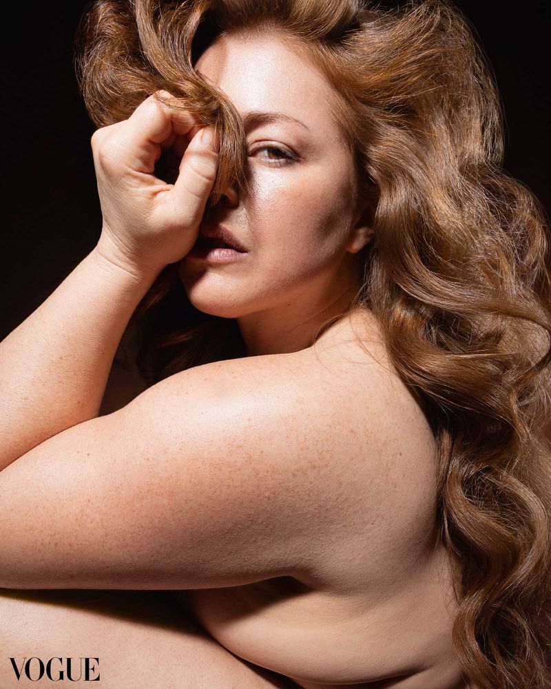 Close-up studio portrait of a bare-shouldered woman with auburn hair and freckled skin, hand near her face, against a dark backdrop.