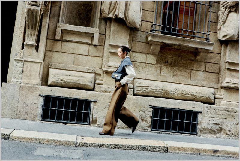 Woman walks on city street in wide-leg pants and vest. Street and architectural details visible as blurred background in daylight.