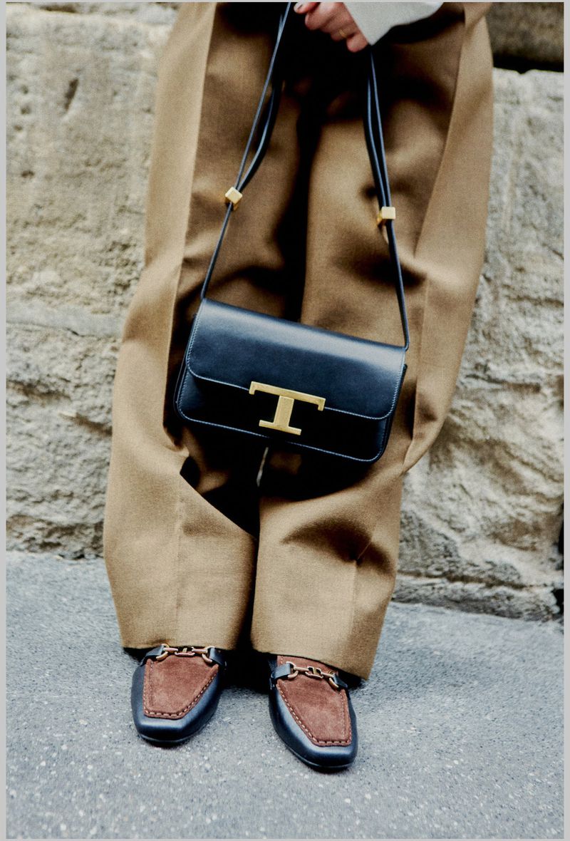 Fashion shoot featuring a black leather Tod's handbag with gold hardware, paired with tan trousers and black and brown Tod's loafers.