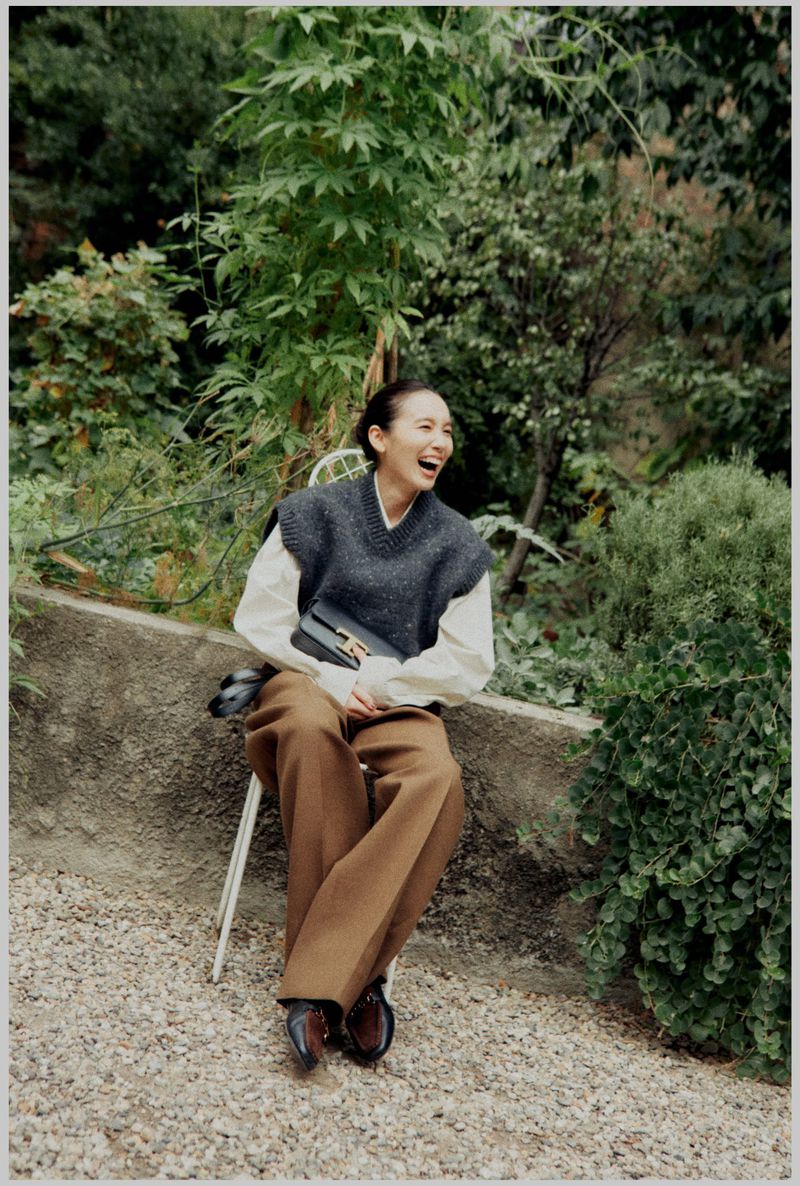 Woman in sweater vest laughs while seated outdoors on a chair, wearing wide-legged brown pants and black loafers.