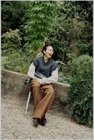 Woman in sweater vest laughs while seated outdoors on a chair, wearing wide-legged brown pants and black loafers.