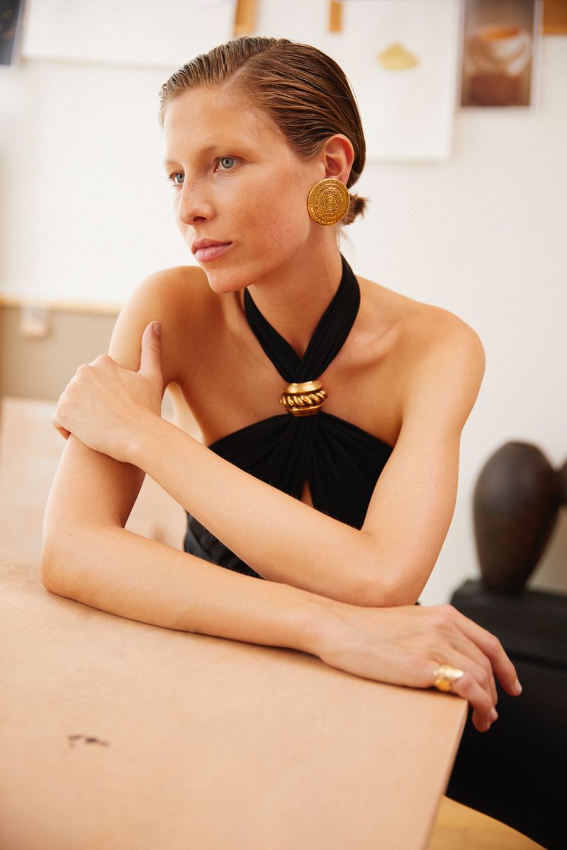 Attractive blonde woman in a black halter top and gold jewelry poses indoors with a neutral expression and arms crossed on a table.