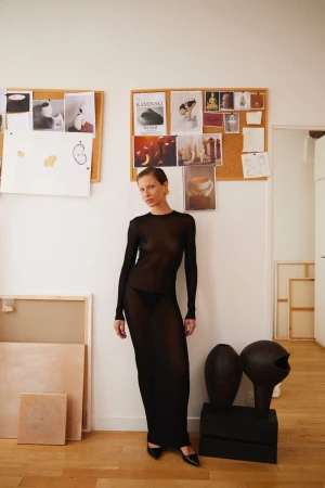 A slender woman in a long sheer black dress poses in an indoor setting with a blonde updo, standing near artwork and decor.