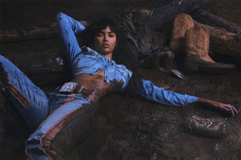 Adult female lying in the mud wearing a denim outfit with boots, a handbag, and a belt buckle with 'USA' text.