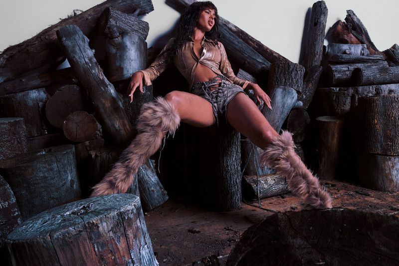 Brunette model poses seated against logs with dramatic light and shadow, wearing shorts and fuzzy boots.