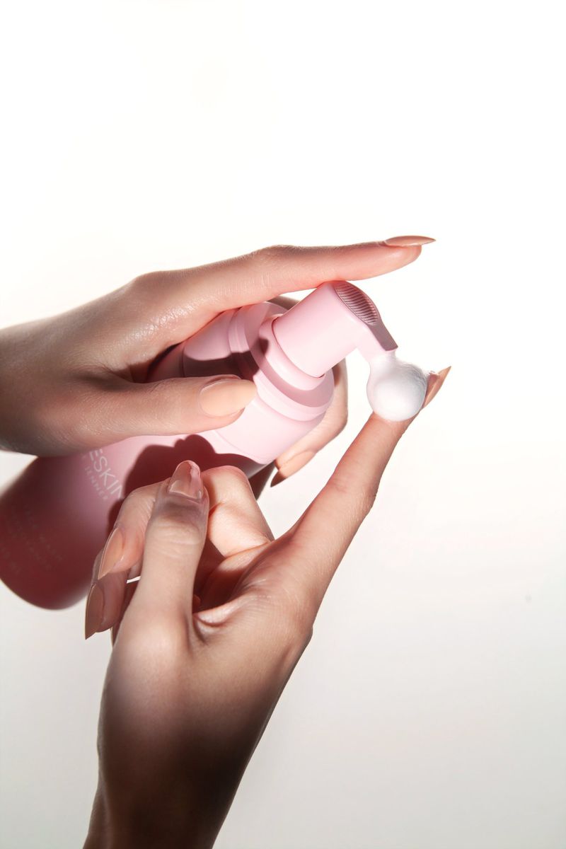Close-up of hands dispensing white foam from a pink bottle, product presentation in a skincare beauty shoot on a white background.