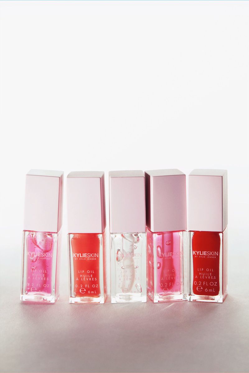 Five Kylie Skin lip oils in pink and clear shades arranged on a white surface, showcasing beauty product presentation.
