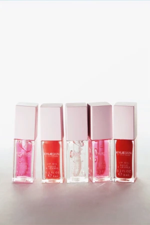 Five Kylie Skin lip oils in pink and clear shades arranged on a white surface, showcasing beauty product presentation.