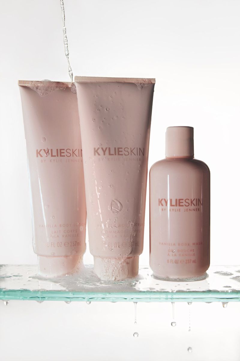 Three light pink Kylie Skin products on a clear glass shelf, with water droplets. A small stream of water pours onto the containers.
