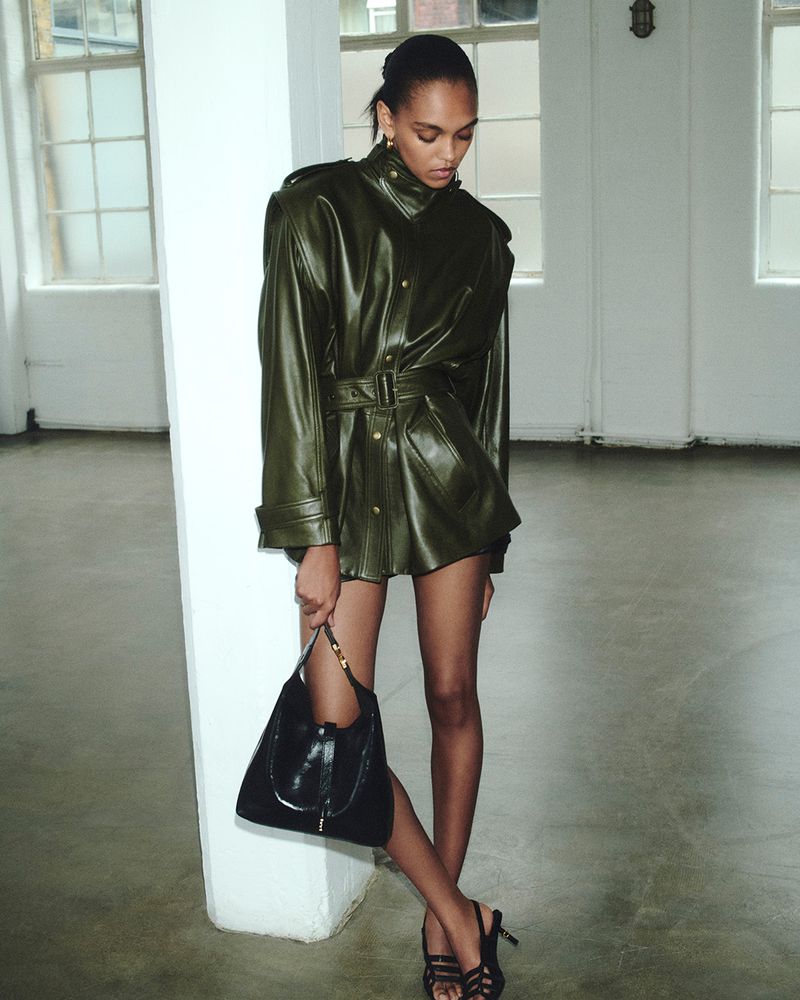 A slender young woman in a belted green leather jacket holds a black purse in an indoor setting with natural light.