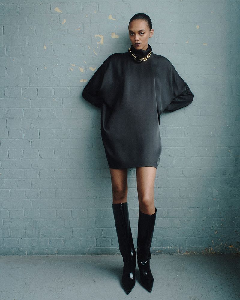 A fashion portrait of a woman in a black mini-dress and black boots standing in front of a blue textured brick wall with a gold chain.