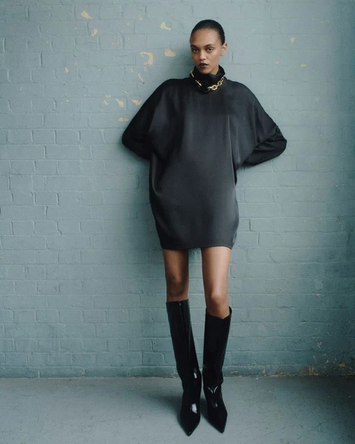 A fashion portrait of a woman in a black mini-dress and black boots standing in front of a blue textured brick wall with a gold chain.