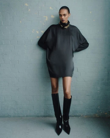 A fashion portrait of a woman in a black mini-dress and black boots standing in front of a blue textured brick wall with a gold chain.