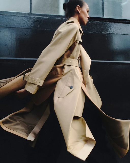 Fashion shot of woman in beige trench coat walking against dark urban wall, side view, showcasing elegance and movement in editorial style.