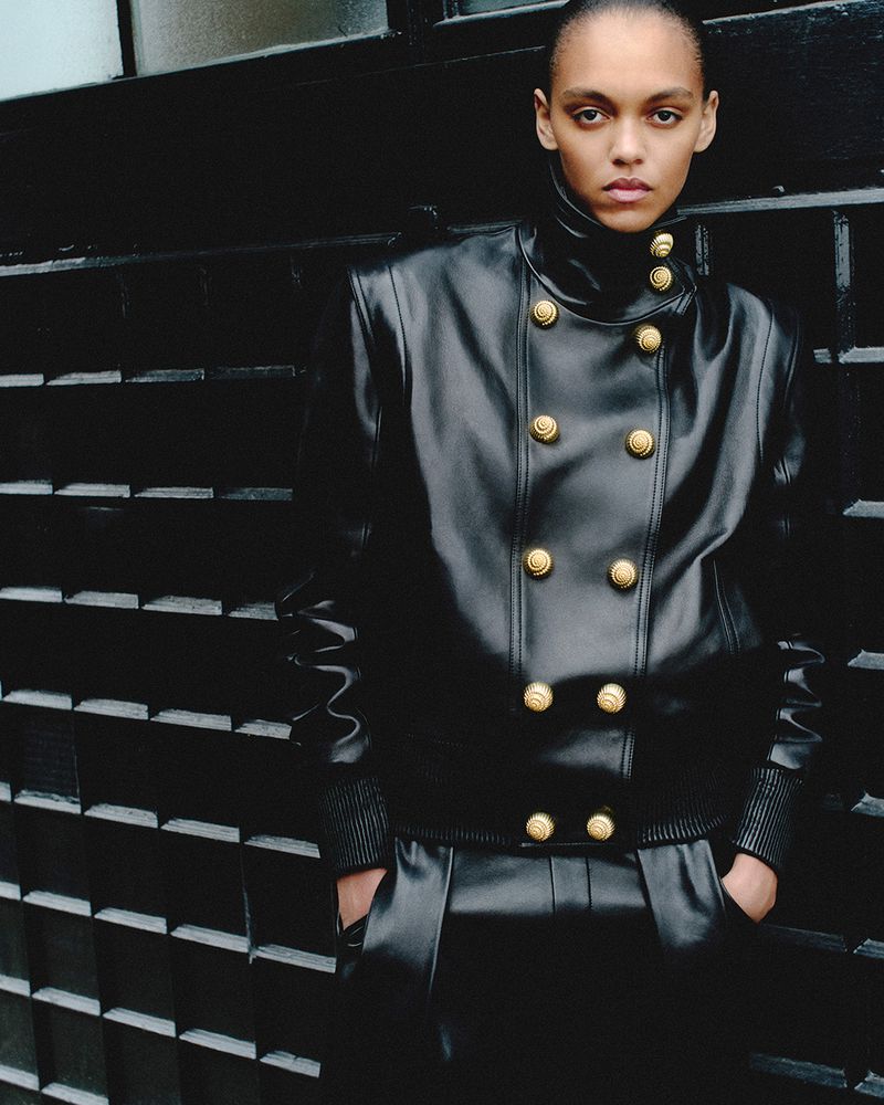 Fashion photo of a woman with dark, short hair wearing a black leather jacket with gold buttons, standing against a black brick wall.