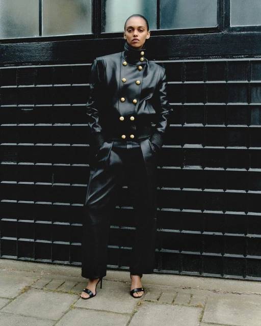 A woman poses outdoors in a black leather jacket and pants with gold buttons, wearing black high-heeled sandals, against a dark wall.
