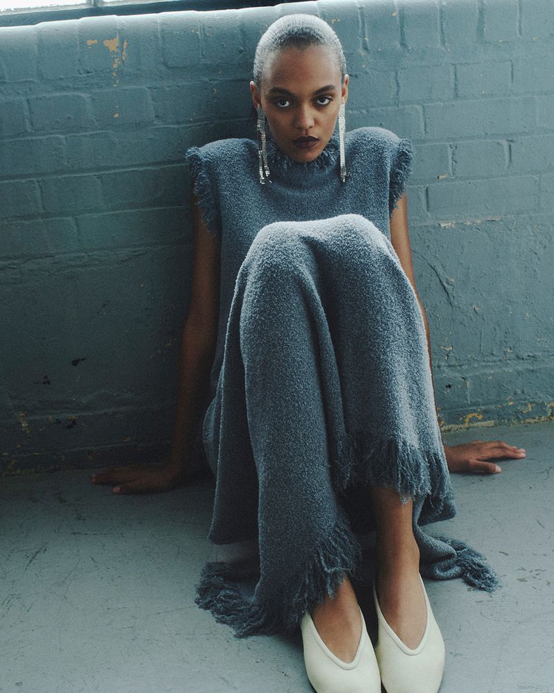 A woman with a shaved head sits on the floor wearing a long gray dress, white shoes, and statement earrings in an indoor setting.