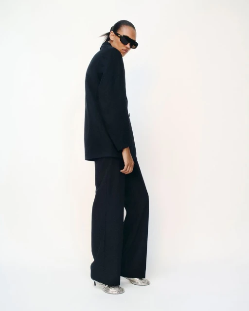 Elegant adult female poses in a black jacket and pants, complemented by silver heels and sunglasses, against a pristine white backdrop.