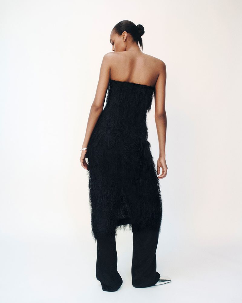 Back view of a black female model in a strapless black dress and trousers. The model has her hair in a bun against a white backdrop.