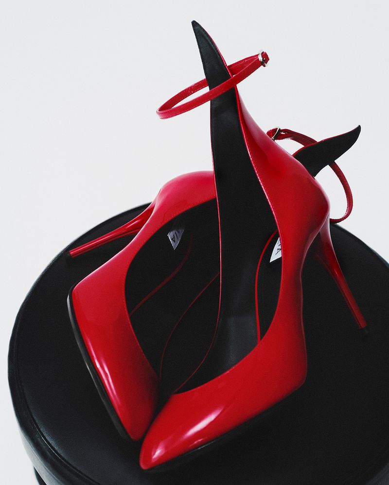 Close-up studio shot of a pair of elegant red patent-leather stiletto high heels with ankle straps sitting on a black stool.