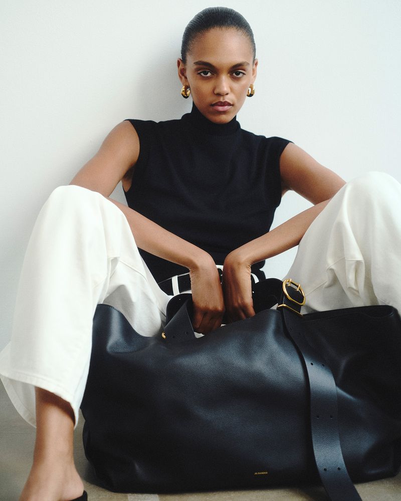 Woman in black top and white pants poses with a black leather tote bag. She is sitting indoors against a white wall.