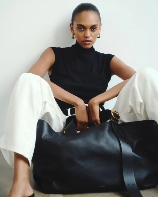 Woman in black top and white pants poses with a black leather tote bag. She is sitting indoors against a white wall.