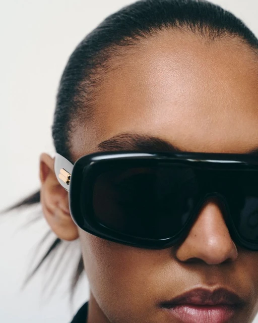 Close-up shot of an adult woman with medium skin tone wearing oversized black sunglasses against a neutral backdrop in a photo studio.