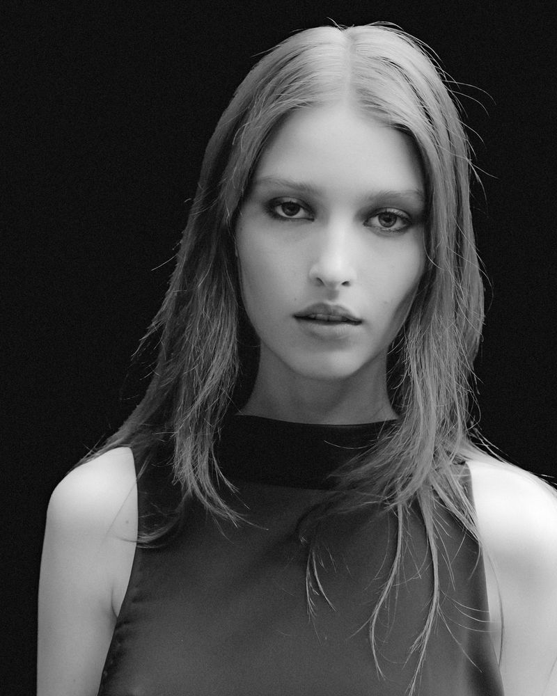 Monochrome portrait of a young adult woman with long hair, wearing a dark sleeveless top, against a black background, looking at camera.