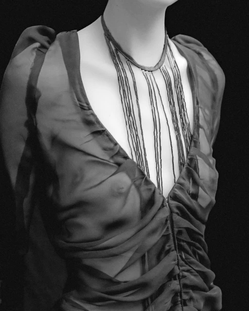 Monochrome image of a mannequin wearing a black sheer dress with a ruched front, accessorized with a beaded necklace against a dark background.