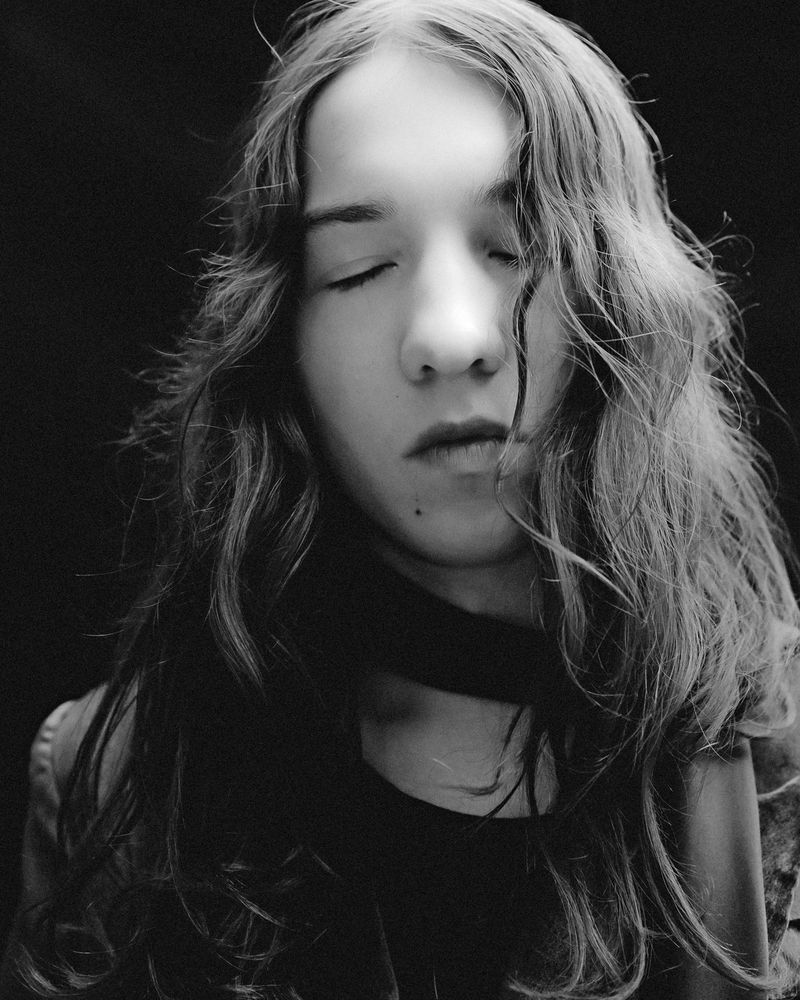 Monochrome portrait of a young adult woman with long wavy hair and closed eyes against a dark background, shot in a studio.