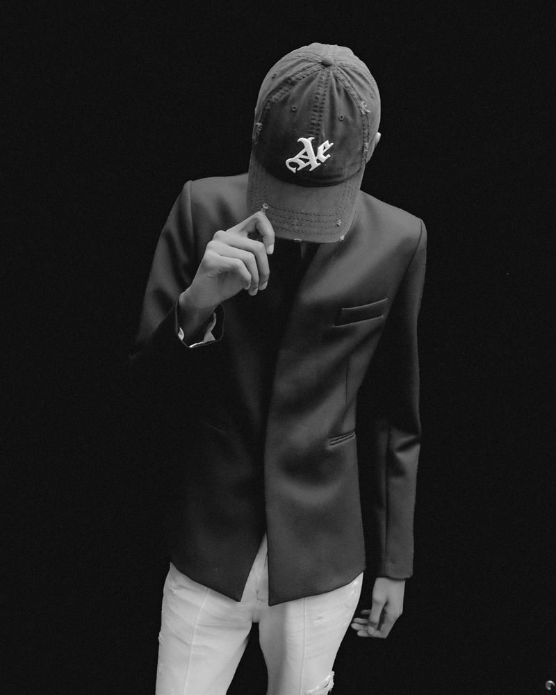 Black and white portrait of an adult male wearing a suit and baseball cap, his face obscured, against a dark background.