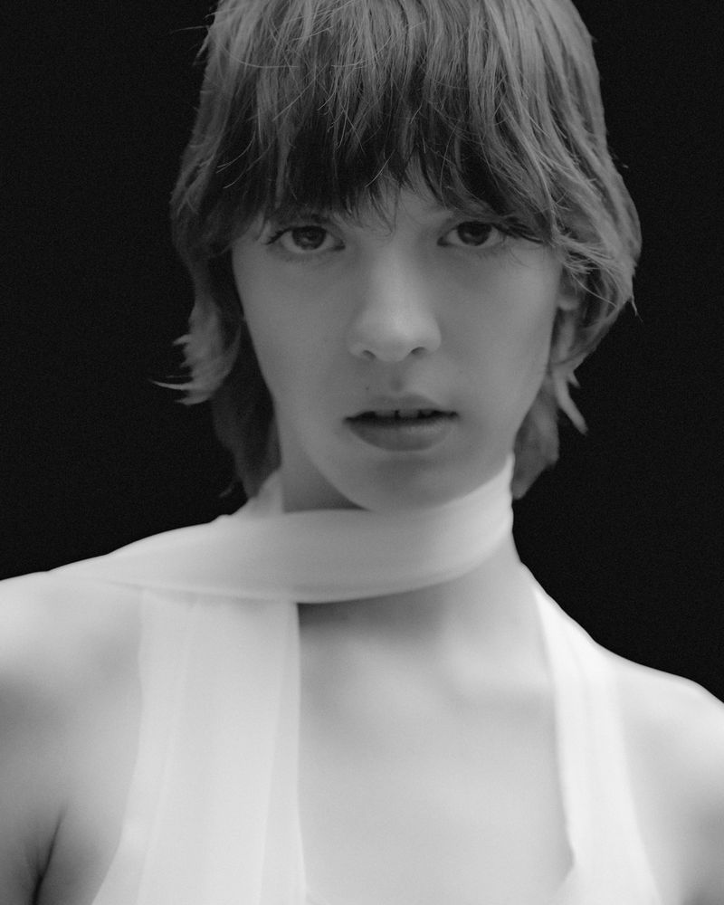 Monochromatic portrait of a young adult with dark hair, light skin, wearing a light-colored scarf, and an ambiguous neutral expression.